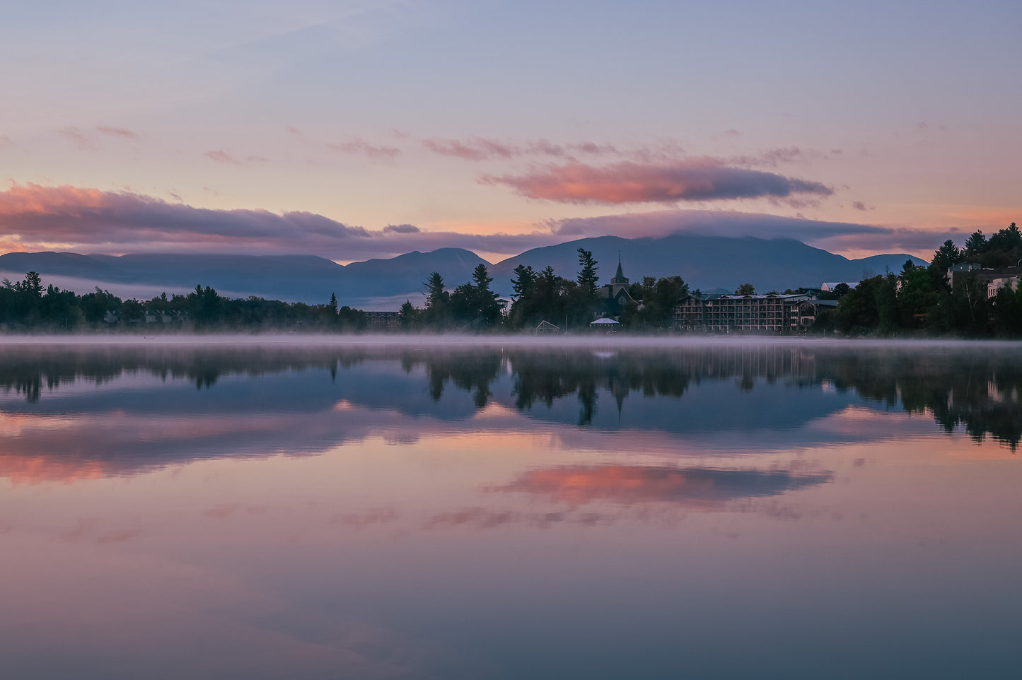 A serene landscape photograph of a calm lake at sunrise or sunset, with mist rising from the water, silhouetted trees, distant mountains, and a soft pink and purple sky reflected in the water.