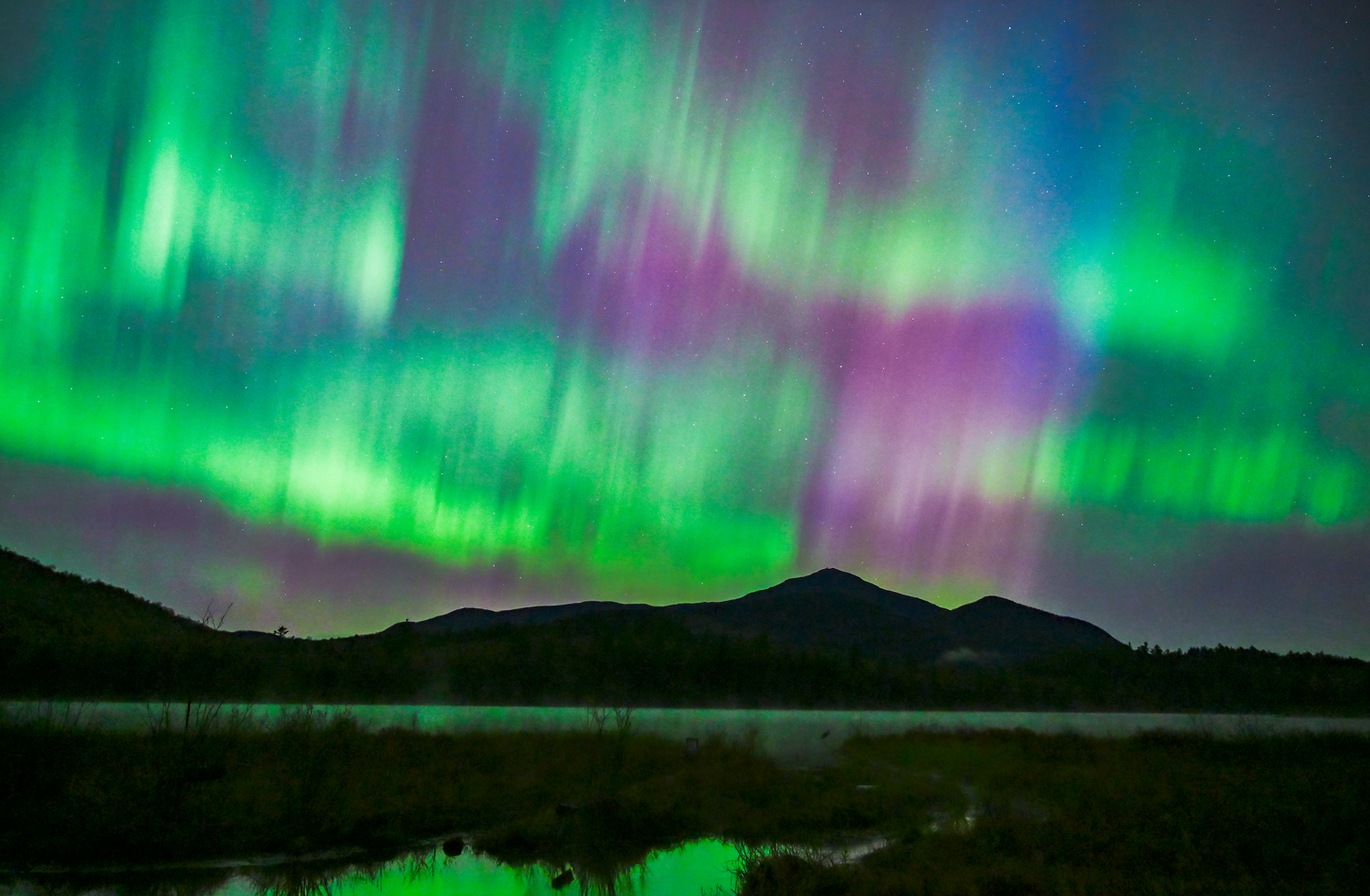 Northern lights with vibrant green, purple, and blue curtains dancing across a night sky above dark mountain silhouettes and a reflective body of water.