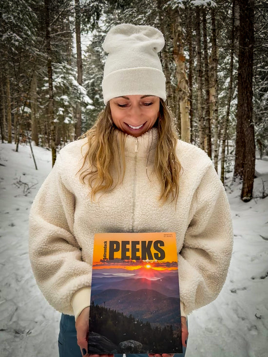 A woman in a snowy forest wearing a cream fleece jacket and matching beanie, holding a copy of the December 2023 issue of Peeks Magazine.