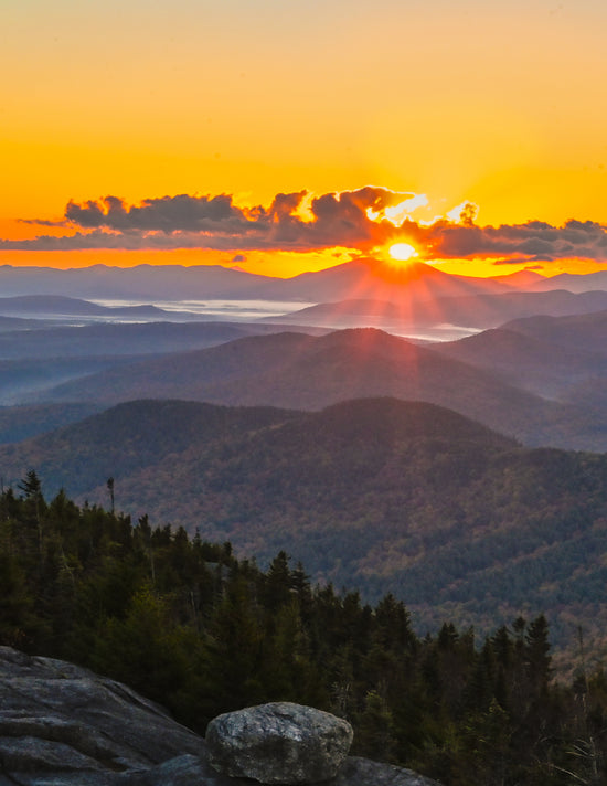 Mountain landscape at sunset with layered forested ridges, a glowing sun near the horizon, and an orange-yellow sky.