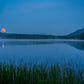A serene blue-toned landscape of a calm lake at dusk, with a glowing orange-red moon near the horizon, dark tree-lined shores, distant mountains, and reeds in the foreground.