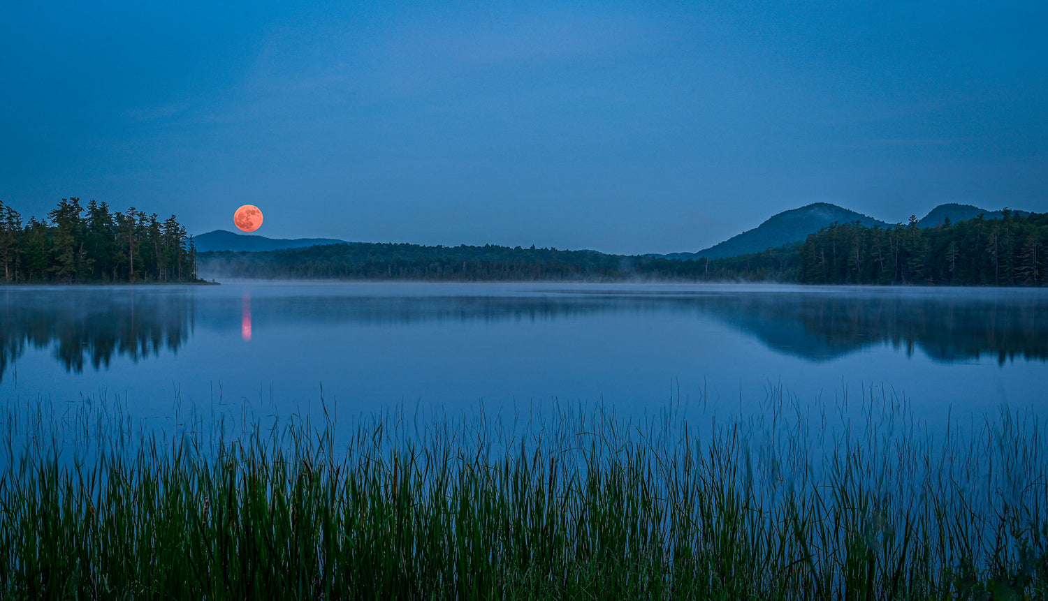 A serene blue-toned landscape of a calm lake at dusk, with a glowing orange-red moon near the horizon, dark tree-lined shores, distant mountains, and reeds in the foreground.