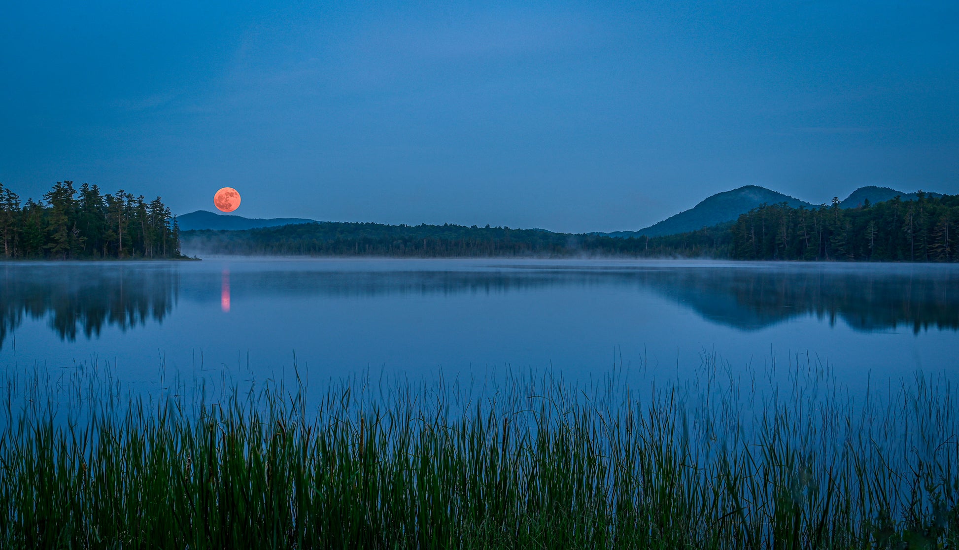 A serene blue-toned landscape of a calm lake at dusk, with a glowing orange-red moon near the horizon, dark tree-lined shores, distant mountains, and reeds in the foreground.