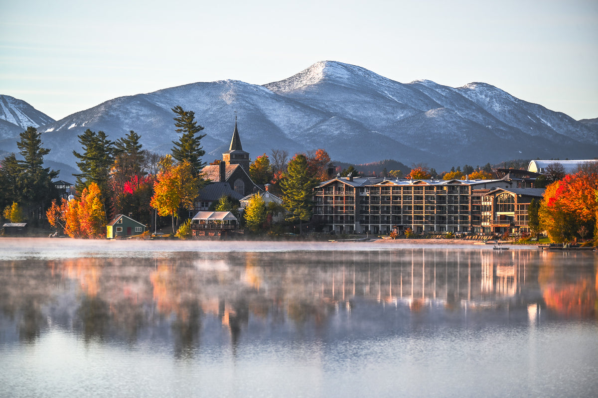 A scenic landscape print showing a mountain resort with a large lodge, a church steeple, and colorful autumn trees reflected in a calm lake under a pale sky.