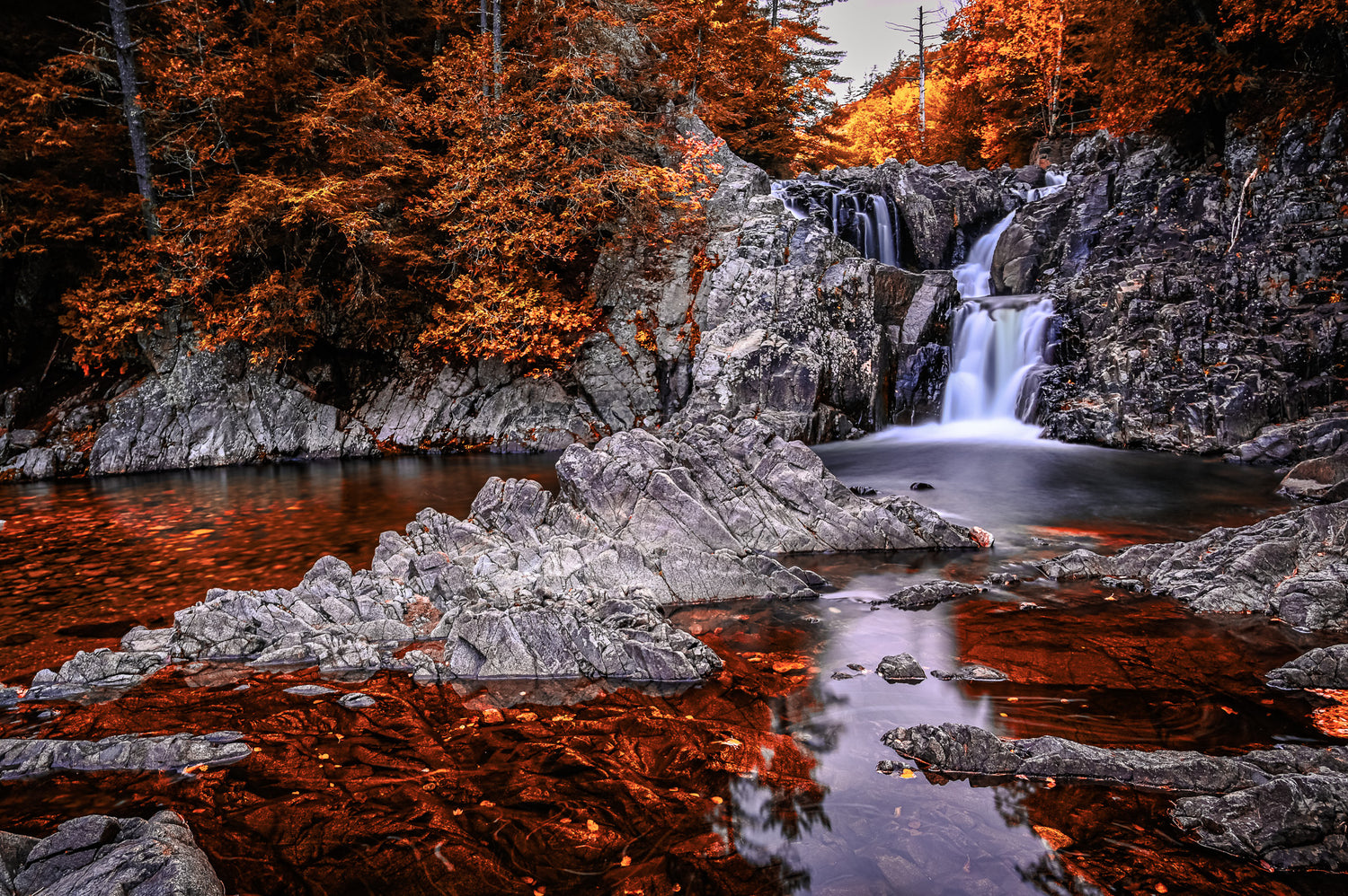 A scenic Adirondack landscape photograph of a waterfall cascading over dark gray rocks into a pool, surrounded by dense autumn foliage in vibrant orange and brown tones.