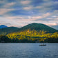 A scenic landscape photograph of a calm lake with a small boat on the water, backed by a forested mountain range and a dramatic cloudy sky at sunset.