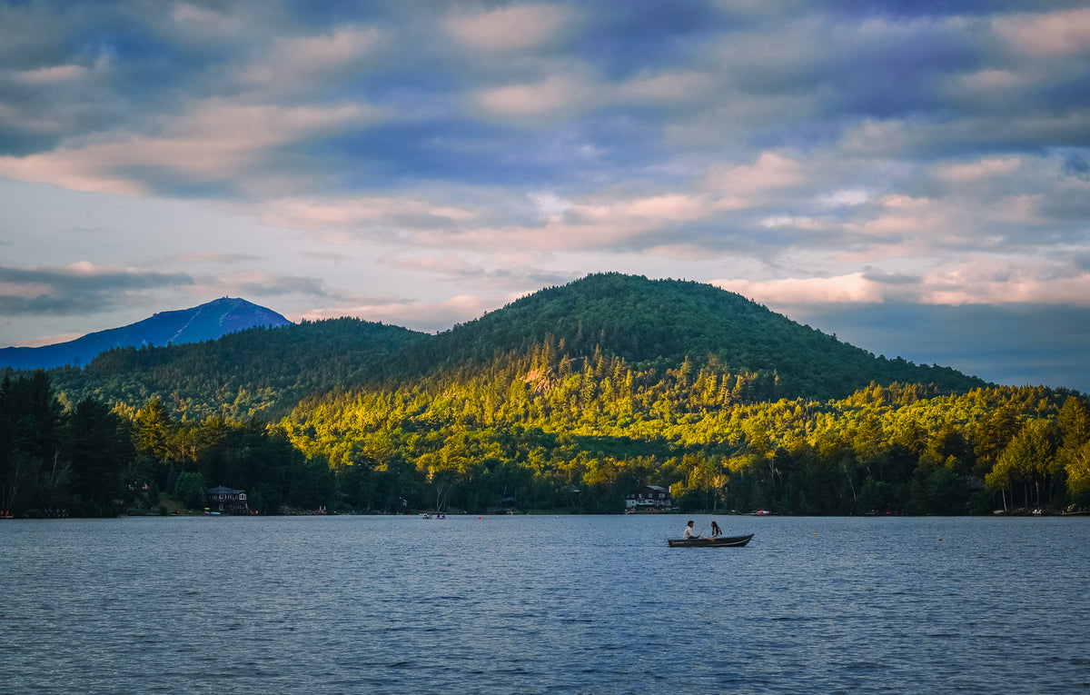 A scenic landscape photograph of a calm lake with a small boat on the water, backed by a forested mountain range and a dramatic cloudy sky at sunset.