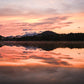 A scenic landscape photograph of a calm lake at sunset, with vibrant orange and pink clouds reflected in the still water, dark tree silhouettes along the horizon, and distant mountains under a warm sky.