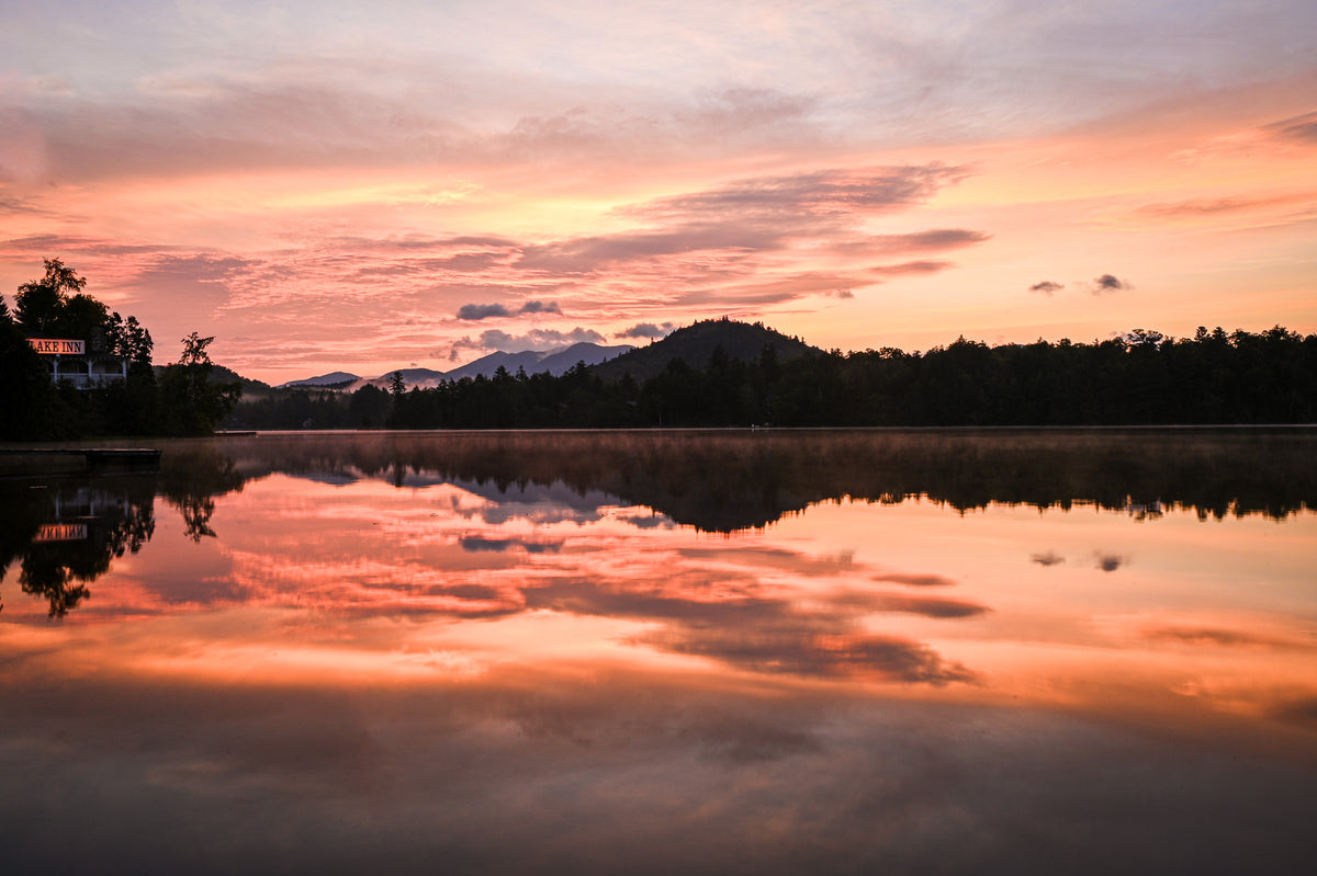 A scenic landscape photograph of a calm lake at sunset, with vibrant orange and pink clouds reflected in the still water, dark tree silhouettes along the horizon, and distant mountains under a warm sky.