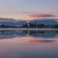 A serene landscape photograph of a calm lake at sunrise or sunset, with mist rising from the water, silhouetted trees, distant mountains, and a soft pink and purple sky reflected in the water.