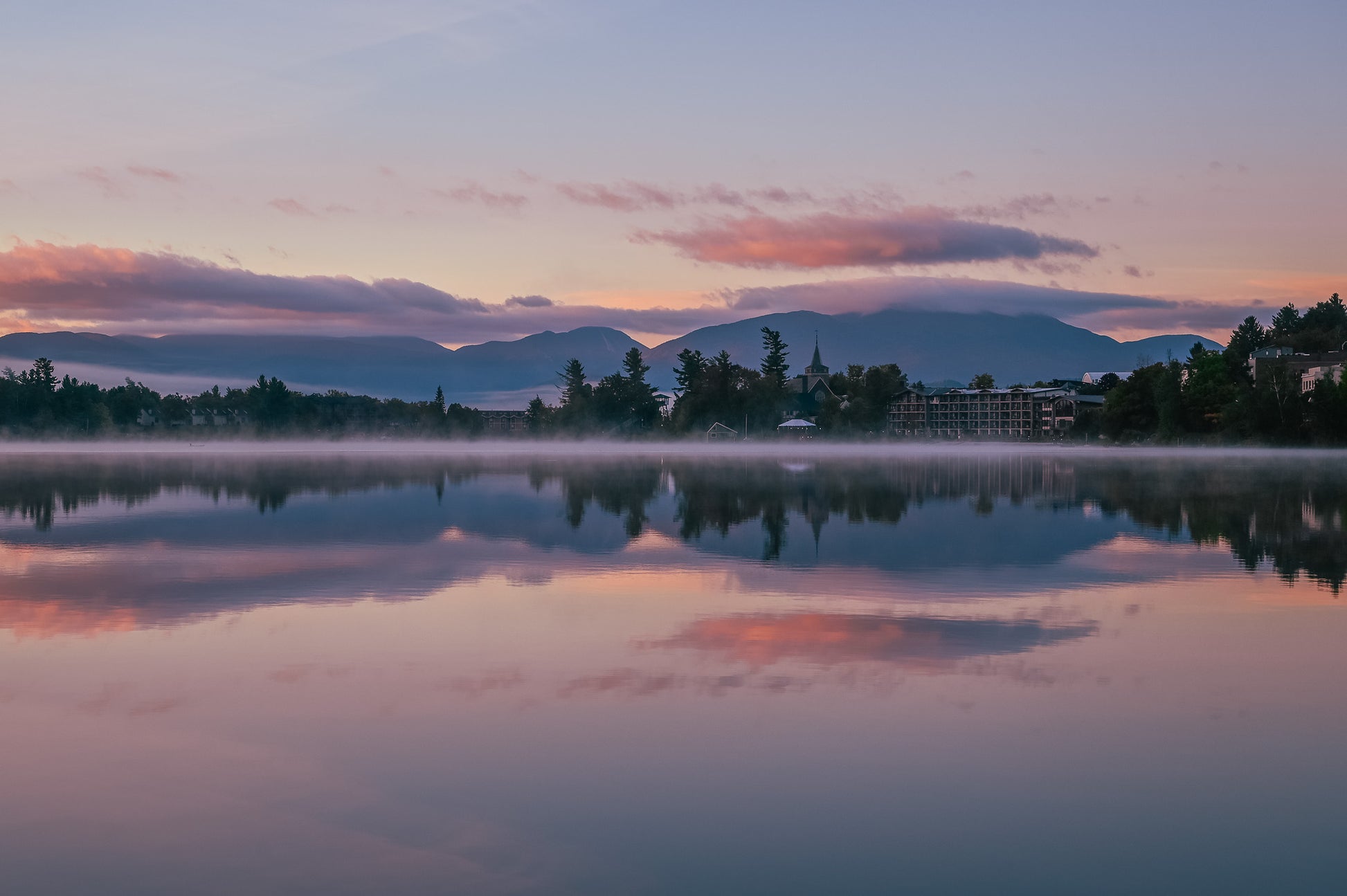 A serene landscape photograph of a calm lake at sunrise or sunset, with mist rising from the water, silhouetted trees, distant mountains, and a soft pink and purple sky reflected in the water.