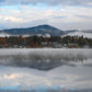A misty mountain lake landscape with autumn trees, small buildings, and a cloudy sky reflected in the calm water.