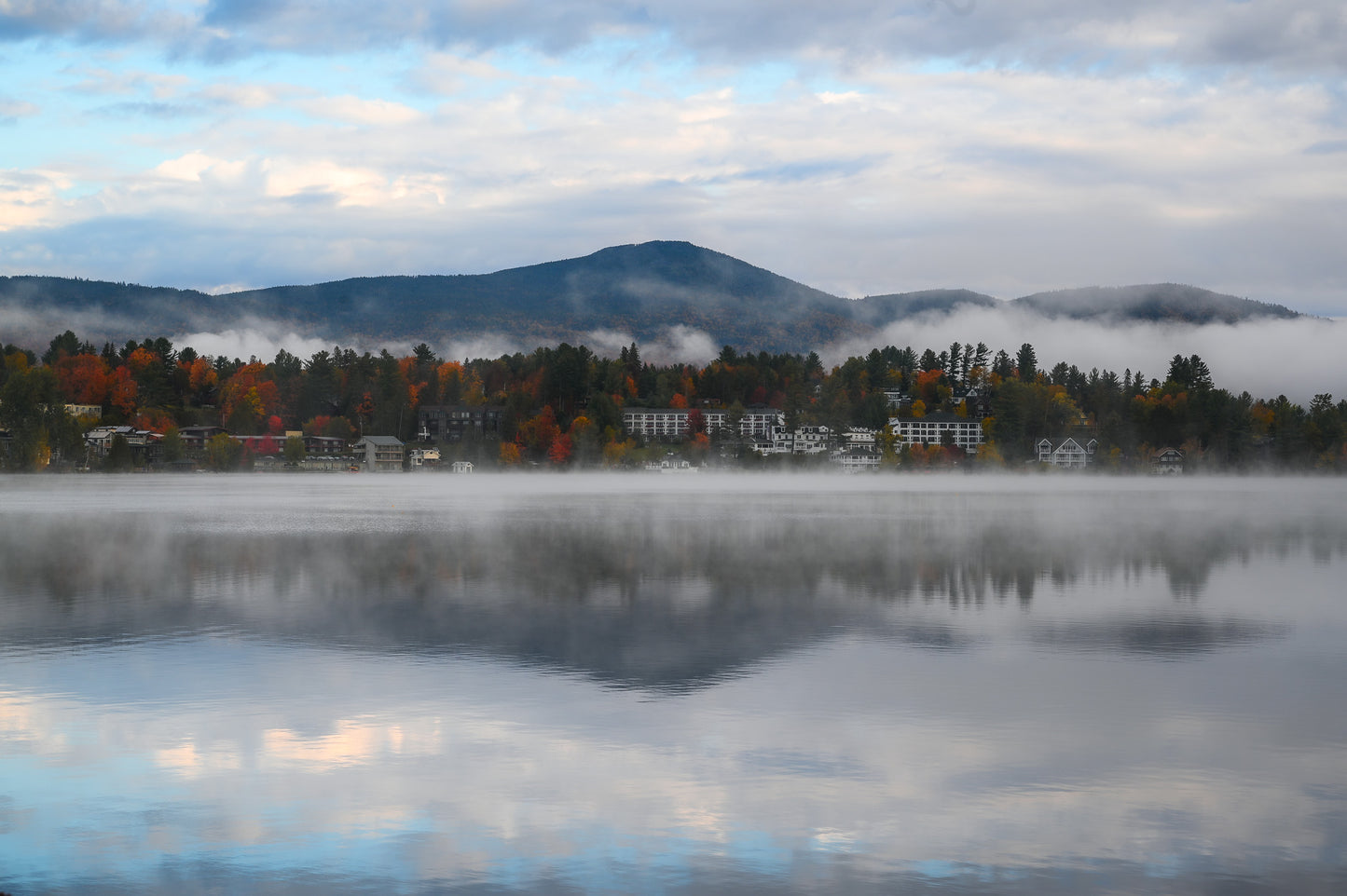 A misty mountain lake landscape with autumn trees, small buildings, and a cloudy sky reflected in the calm water.