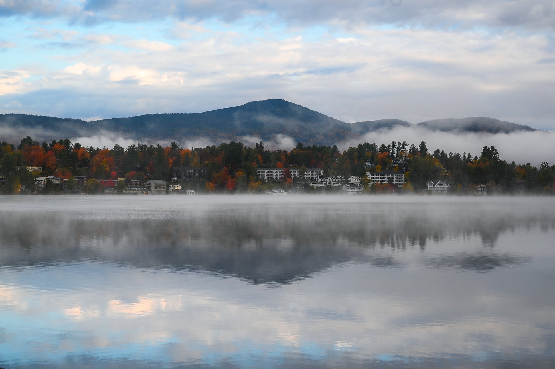 A misty mountain lake landscape with autumn trees, small buildings, and a cloudy sky reflected in the calm water.