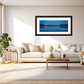 A framed landscape art print depicting a serene blue lake at dusk, hung on a light beige wall above a modern beige sofa in a bright, minimalist living room.