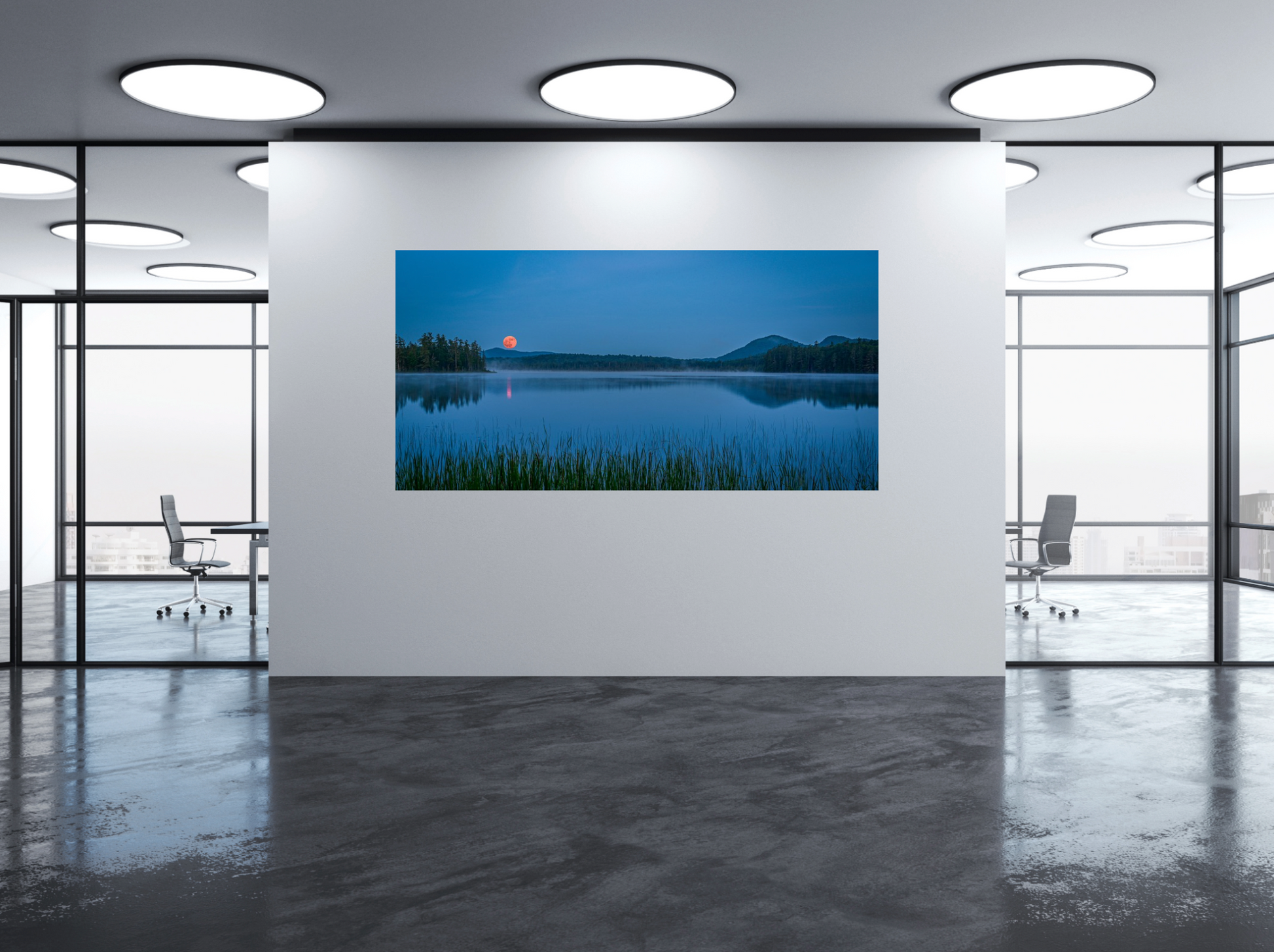 A framed or frameless landscape wall art print showing a calm lake at dusk with blue sky, dark tree silhouettes, and a moon near the horizon, displayed on a white wall in a modern office interior with glass walls and ceiling lights.