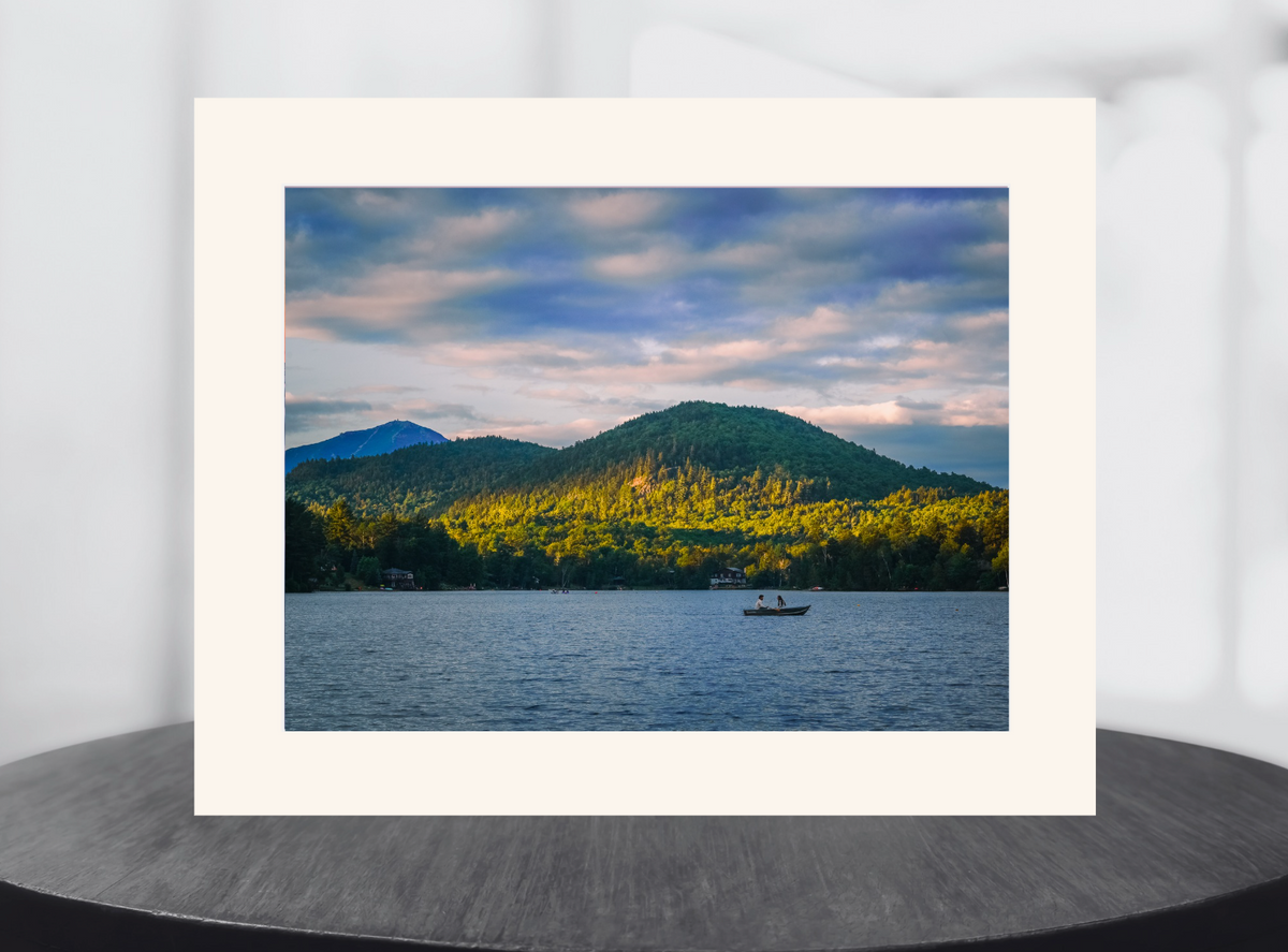 landscape art print depicting a lake with green mountains and a small boat under a cloudy sky, displayed in a white matte border.