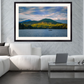 A framed landscape art print depicting a lake with green mountains and a small boat under a cloudy sky, displayed on a gray marble wall above a modern white sectional sofa in a living room setting.