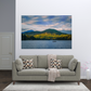 A framed landscape wall art print showing a mountain range over a lake with a small boat, displayed above a light gray sofa in a modern living room setting.