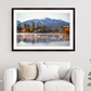 A framed landscape art print of a mountain lake with autumn trees and a building, displayed on a light gray wall above a white sofa in a styled living room.