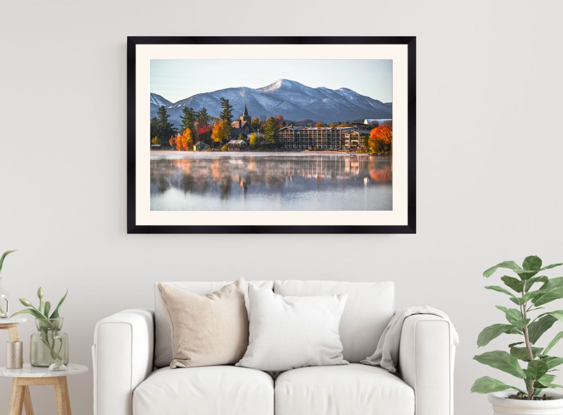 A framed landscape art print of a mountain lake with autumn trees and a building, displayed on a light gray wall above a white sofa in a styled living room.