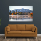 A framed wall art print showing a mirror lake scene with autumn trees and a resort building, displayed above a mustard-brown sofa in a styled living room.