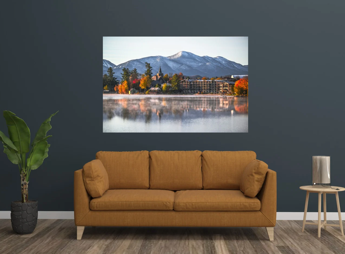 A framed wall art print showing a mirror lake scene with autumn trees and a resort building, displayed above a mustard-brown sofa in a styled living room.