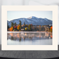 A framed art print featuring a scenic mountain landscape with autumn trees, a church steeple, and a lake reflection, displayed on a dark round table against a light gray background.