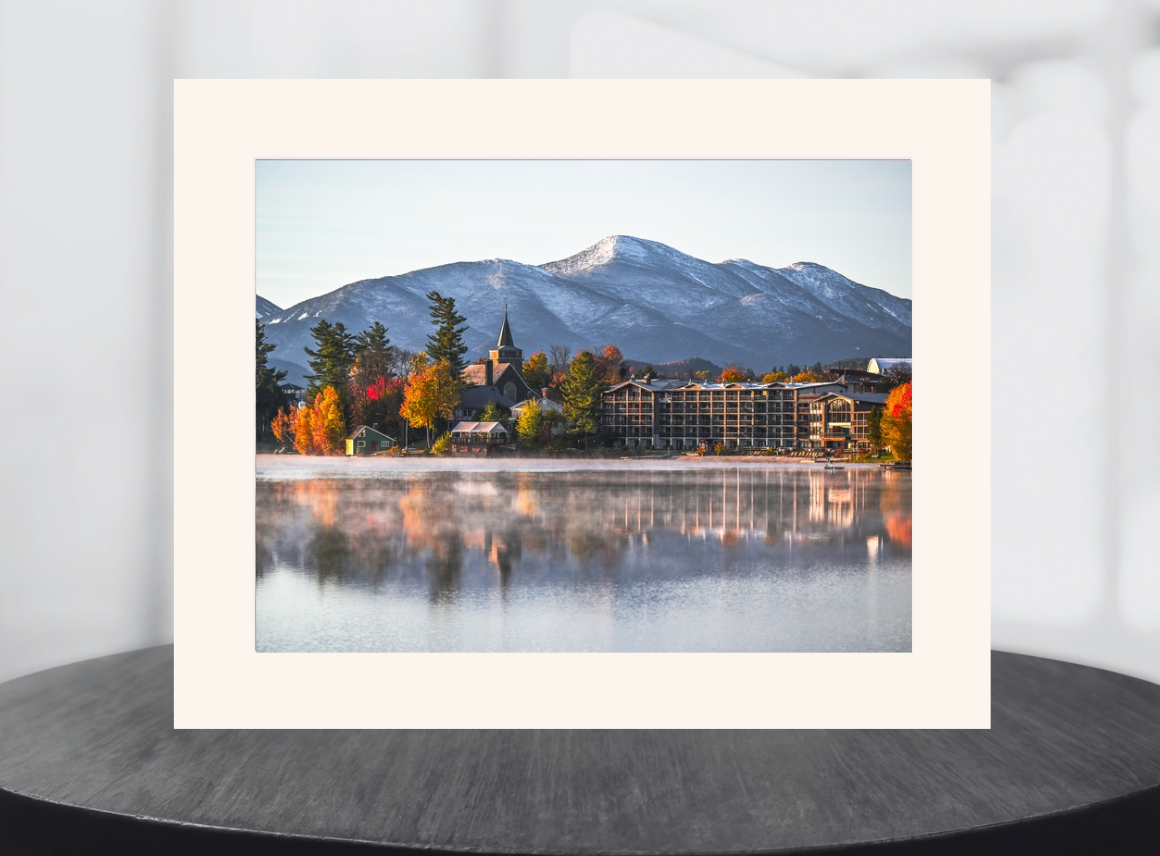 A framed art print featuring a scenic mountain landscape with autumn trees, a church steeple, and a lake reflection, displayed on a dark round table against a light gray background.