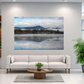 A large rectangular wall art print showing a misty mountain lake landscape with blue sky and reflections, displayed above a white sofa in a modern living room with potted plants.