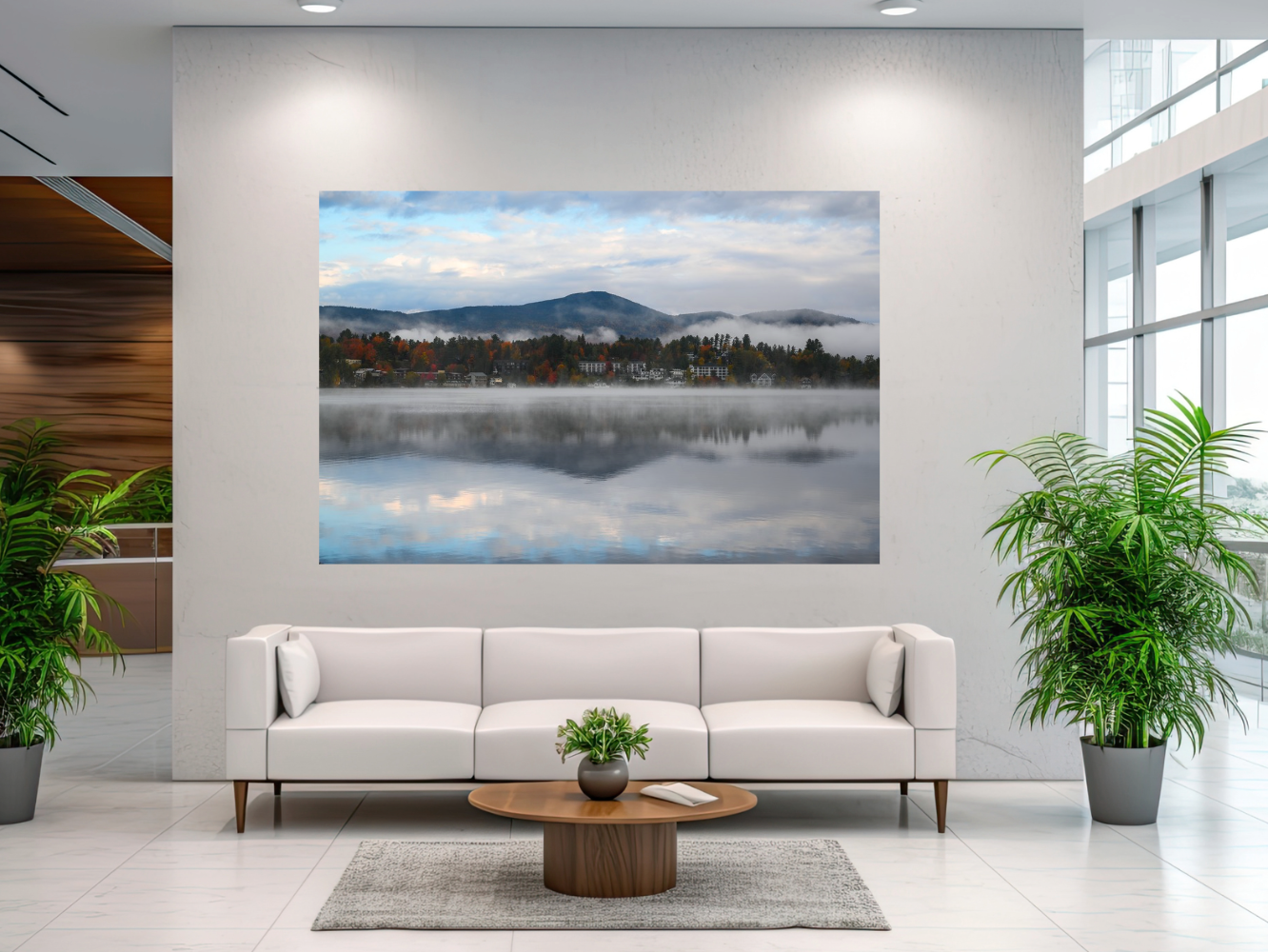 A large rectangular wall art print showing a misty mountain lake landscape with blue sky and reflections, displayed above a white sofa in a modern living room with potted plants.