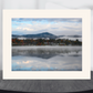A matted landscape art print featuring a misty mountain lake scene with autumn trees and a small village, displayed on a dark round table against a light gray background.
