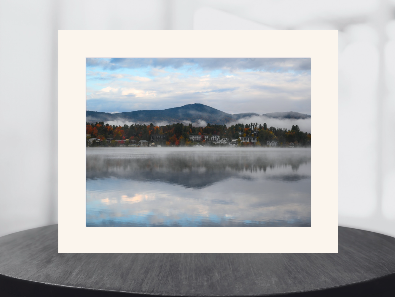 A matted landscape art print featuring a misty mountain lake scene with autumn trees and a small village, displayed on a dark round table against a light gray background.