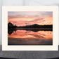 A matted art print featuring a scenic sunset landscape with pink, orange, and purple clouds reflected in calm water, displayed on a dark round surface against a light gray background.