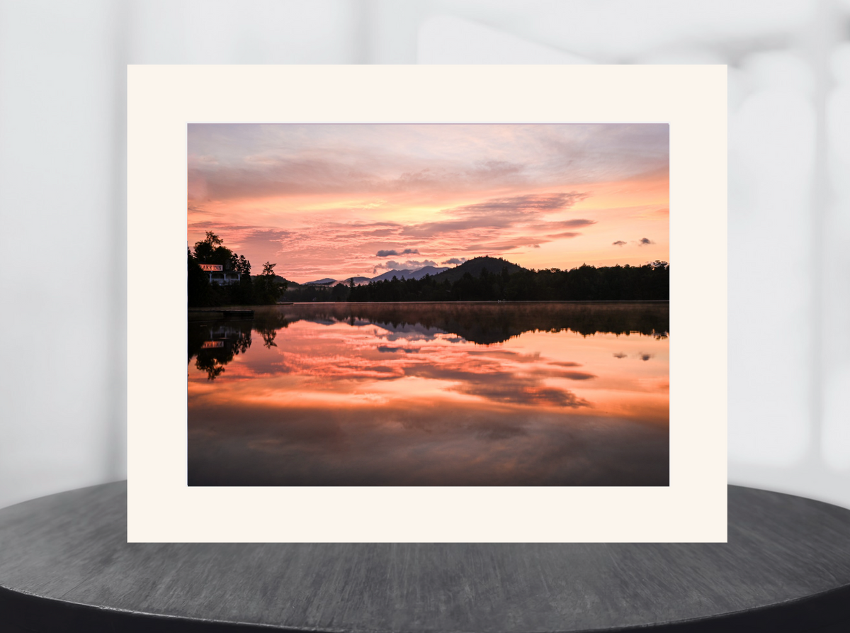 A matted art print featuring a scenic sunset landscape with pink, orange, and purple clouds reflected in calm water, displayed on a dark round surface against a light gray background.