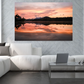 A large rectangular wall art print showing a colorful sunset over a reflective lake, displayed above a white sectional sofa in a modern living room setting.