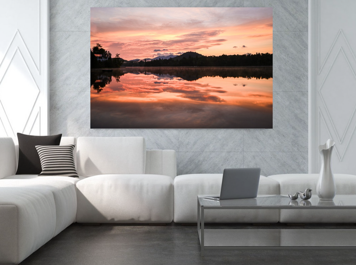A large rectangular wall art print showing a colorful sunset over a reflective lake, displayed above a white sectional sofa in a modern living room setting.
