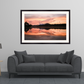 A framed landscape art print of a sunset over a lake reflection, displayed on a light gray wall above a modern gray sofa in a styled living room setting.