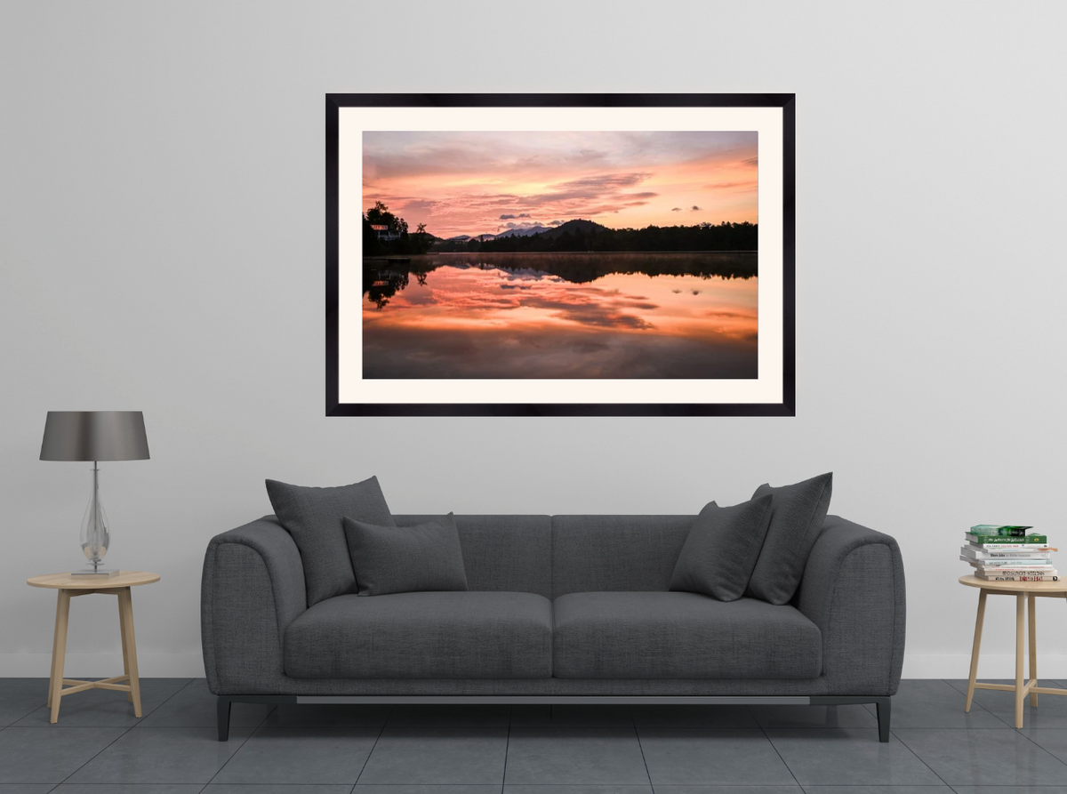 A framed landscape art print of a sunset over a lake reflection, displayed on a light gray wall above a modern gray sofa in a styled living room setting.