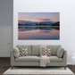 A large rectangular wall art print showing a calm lake reflection at sunset, displayed above a light gray sofa in a modern living room setting.
