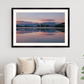 A framed landscape art print of a serene lake sunset with pink and purple clouds reflected in the water, displayed on a white wall above a modern white sofa with decorative pillows.