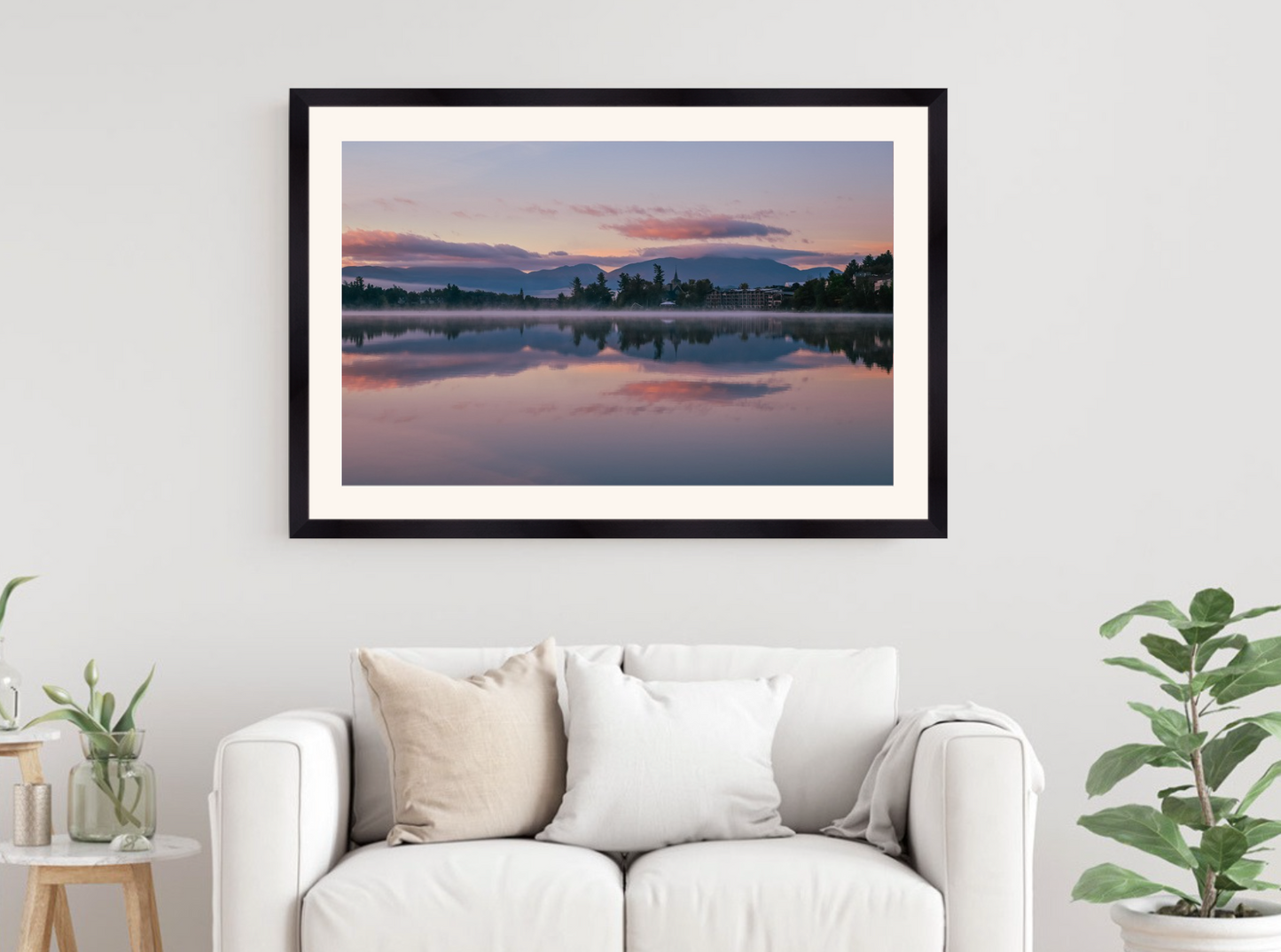 A framed landscape art print of a serene lake sunset with pink and purple clouds reflected in the water, displayed on a white wall above a modern white sofa with decorative pillows.
