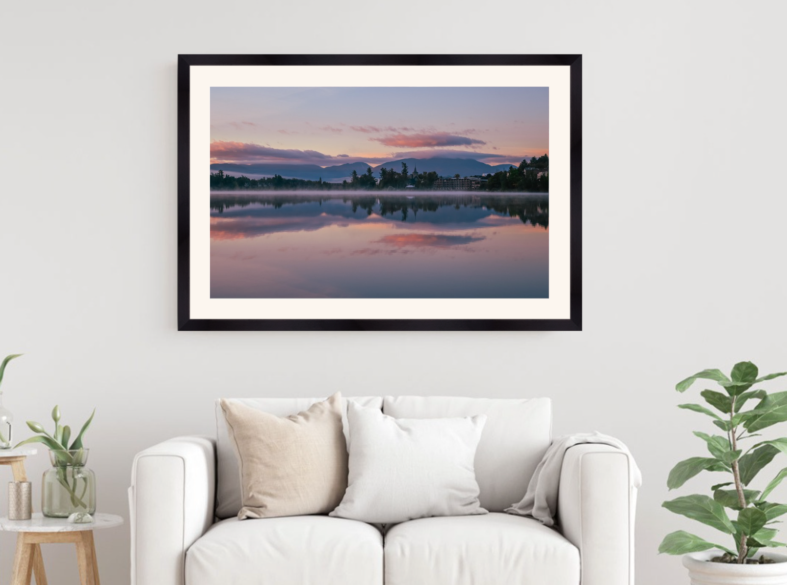 A framed landscape art print of a serene lake sunset with pink and purple clouds reflected in the water, displayed on a white wall above a modern white sofa with decorative pillows.